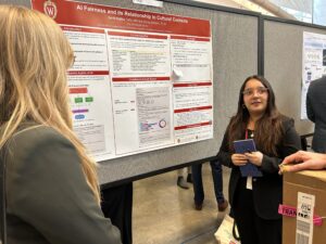 Emili Robles wearing glasses and holding a notepad as she presents her work shown on a poster behind her. 