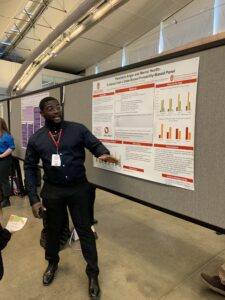 Jeremyah Williams wearing a black button-up shirt, presenting the findings on his poster to onlookers.
